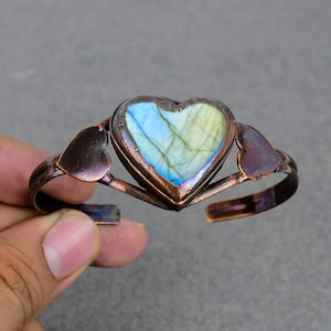 May include: A copper cuff bracelet with a heart-shaped labradorite gemstone. The bracelet is made of copper wire and has a hammered finish. The labradorite gemstone is iridescent and has a blue and green color.