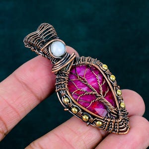 May include: A wire-wrapped pendant featuring a teardrop-shaped magenta gemstone with an integrated copper wire tree design. The pendant includes a small round white moonstone at the top and is accented with tiny gold-toned metal beads along the edges.
