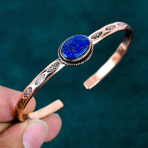 May include: A copper cuff bracelet featuring an oval-shaped, deep blue lapis lazuli gemstone set in a twisted metal border. The band is decorated with repeating geometric patterns etched into the metal surface, creating a textured appearance.