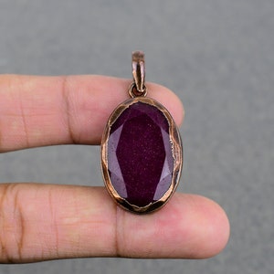 Puede incluir: Un colgante de cobre con una piedra preciosa de color púrpura oscuro engastada en una forma ovalada facetada.