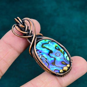 May include: An oval pendant featuring an iridescent abalone shell inlay with swirling blue, green, and purple tones. The shell is encased in dark, oxidized copper wire wrapping with intricate coils and three small brass beads at the base.