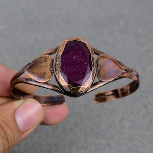 May include: A copper cuff bracelet with a large oval-shaped purple gemstone set in the center. The bracelet is decorated with two heart-shaped cutouts on either side of the gemstone.