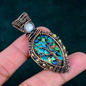 May include: A teardrop-shaped pendant featuring an iridescent abalone shell inlay with a copper wire-wrapped tree design. The pendant is accented with small gold-toned beads and a round, translucent white moonstone set at the top bail.