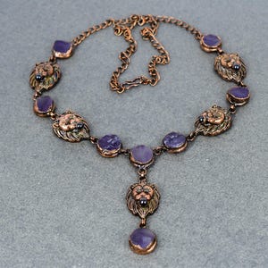 Pode incluir: Um colar em tom de cobre com pingentes de cabeça de leão e detalhes em pedras preciosas roxas. O colar tem um design de corrente com vários pingentes de cabeça de leão e pedras roxas redondas. O pingente tem uma única cabeça de leão.
