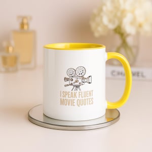 May include: A white ceramic mug with a bright yellow interior and handle. The front features a black line drawing of a vintage film camera above the text "I SPEAK FLUENT MOVIE QUOTES" printed in a tan, sans-serif font on the white surface.