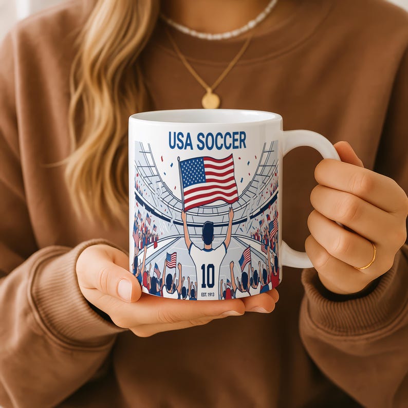 May include: White ceramic mug with a wraparound design featuring a soccer stadium scene. The design includes the text "USA SOCCER", a player holding a U.S. flag, and the number "10". The mug has a curved handle.