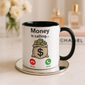 May include: White ceramic mug with a black interior and handle, featuring the text "Money is calling..." and a graphic of a money bag with a dollar sign. The mug also has "Decline" and "Accept" icons.