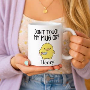以下が含まれることがあります： 「DON'T TOUCH MY MUG OK？」の文字とナイフを持った黄色いひよこの漫画が描かれた白いセラミックマグカップ。「Henry」という名前が下に印刷されています。マグカップには白いハンドルが付いています。