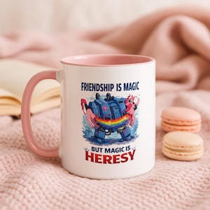 Può includere: Una tazza in ceramica bianca con manico e bordo interno rosa. La parte anteriore presenta un grafico di un robot blu e un pony rosa con un arcobaleno, accompagnato dal testo: Friendship is magic but magic is heresy.