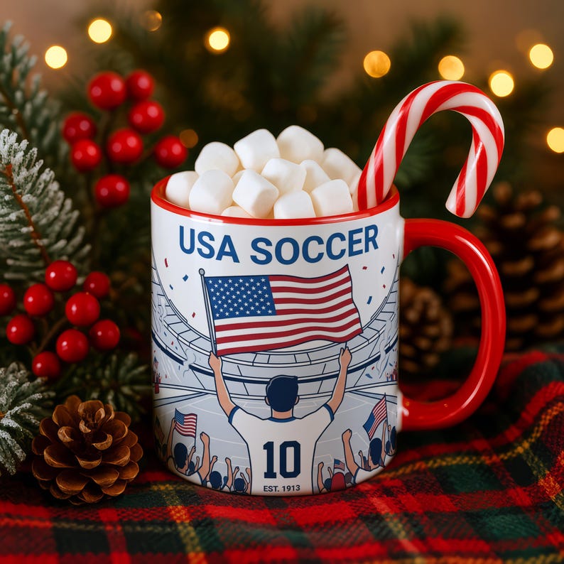 May include: A white ceramic mug with a red handle and rim, filled with marshmallows and a candy cane. The mug features a graphic of a soccer player holding a US flag, with the text "USA SOCCER" and "10 EST. 1913".