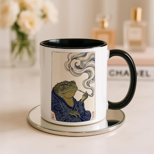 May include: White ceramic mug with a black interior and handle. The mug features an illustration of a green frog in a blue robe smoking a pipe, with stylized smoke. The mug sits on a silver coaster.