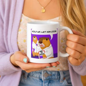 Op de afbeelding: Witte keramische mok met een cartoon marmot in een labjas, met een klembord met het opschrift "Lab Protocol". De mok heeft de tekst "Hold up, let him cook". De marmot staat tegen een paarse achtergrond.