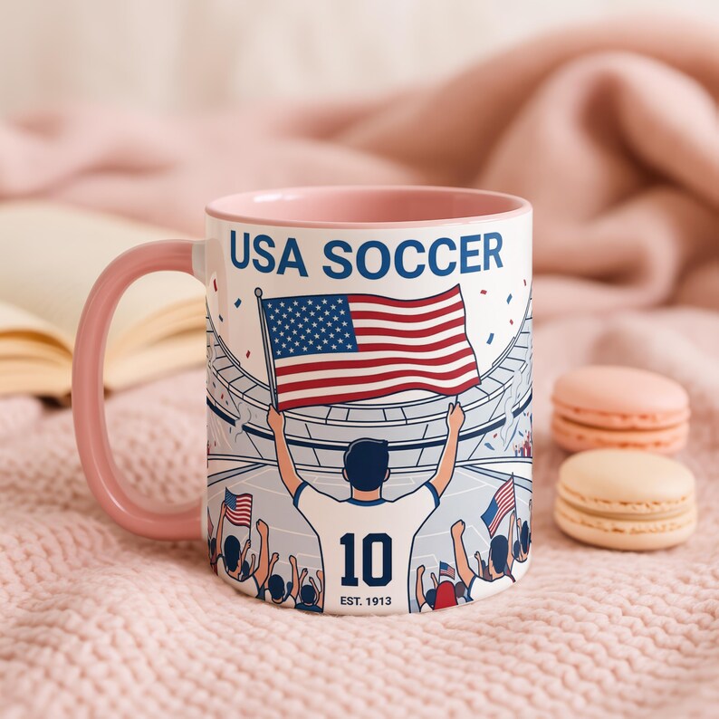 May include: A white ceramic mug with a pink handle and interior. The mug features a graphic of a soccer player holding an American flag in a stadium, with the text "USA SOCCER" and "10 EST. 1913".