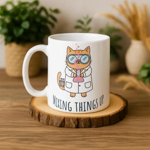 Può includere: Tazza in ceramica bianca con un gatto dei cartoni animati con un camice da laboratorio e occhiali, che tiene in mano un pallone. La tazza ha la scritta "MIXING THINGS UP" e poggia su un sottobicchiere di legno. Il gatto è arancione e bianco.