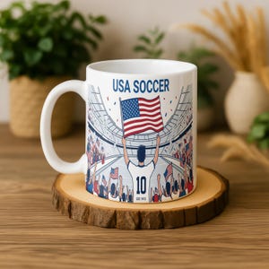 May include: White ceramic mug featuring a soccer stadium scene with the text "USA SOCCER." The design includes a player holding a US flag, the number 10, and the text "EST. 1913." The mug sits on a wooden coaster.
