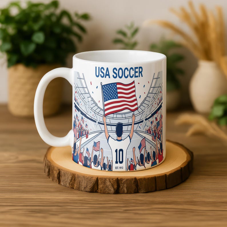 May include: White ceramic mug featuring a soccer stadium scene with the text "USA SOCCER." The design includes a player holding a US flag, the number 10, and the text "EST. 1913." The mug sits on a wooden coaster.