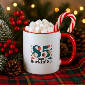 Puede incluir: Una taza de cerámica blanca con borde y asa rojos, llena de malvaviscos blancos y un bastón de caramelo rojo y blanco. El frente presenta el texto 85 y Still Rockin' 85 en verde oscuro y naranja, rodeado de formas geométricas.
