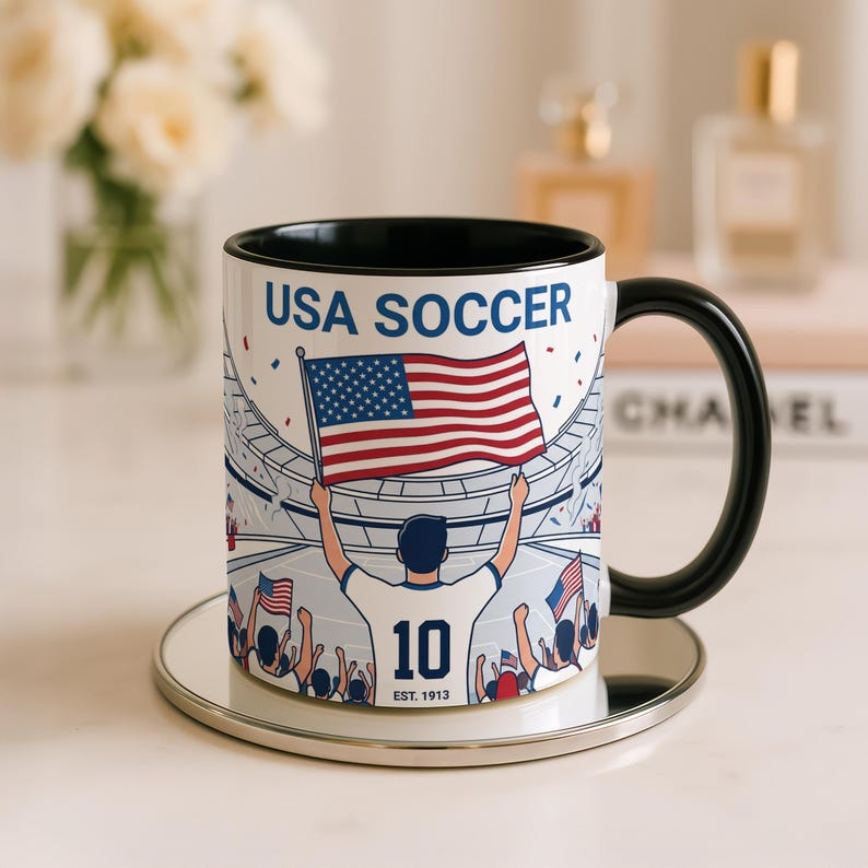 May include: White ceramic mug with a black handle and rim, featuring a USA Soccer design. The design includes a player holding an American flag, the text "USA SOCCER", and the number "10". The mug is on a silver coaster.