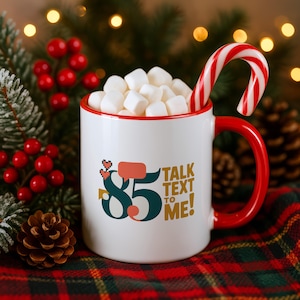 May include: A white ceramic mug with a red rim and handle, filled with white marshmallows and a red and white candy cane. The mug features the dark teal number 85, a red speech bubble, and the gold text TALK TEXT TO ME! printed on the front.