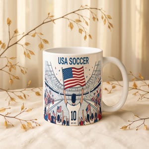 May include: White ceramic mug featuring a USA Soccer design. The graphic depicts a soccer player holding an American flag in a stadium filled with cheering fans. The text "USA SOCCER" is printed at the top.