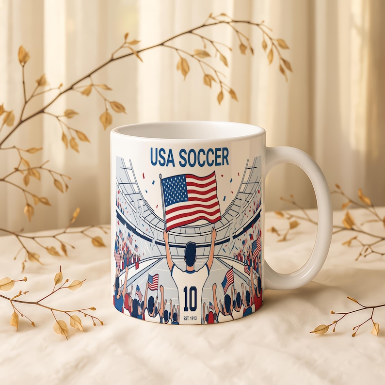 May include: White ceramic mug featuring a USA Soccer design. The graphic depicts a soccer player holding an American flag in a stadium filled with cheering fans. The text "USA SOCCER" is printed at the top.