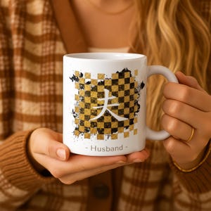 May include: White ceramic mug with a gold and black checkered pattern, a Japanese character, and the word "Husband". The mug has a white handle. The mug is held by a person.