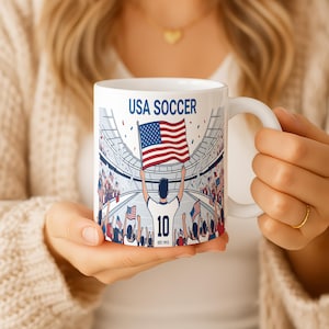 May include: White ceramic mug featuring a USA Soccer design. The mug displays the text "USA SOCCER" above an illustration of a soccer player holding an American flag in a stadium filled with fans. The player's jersey number is 10.