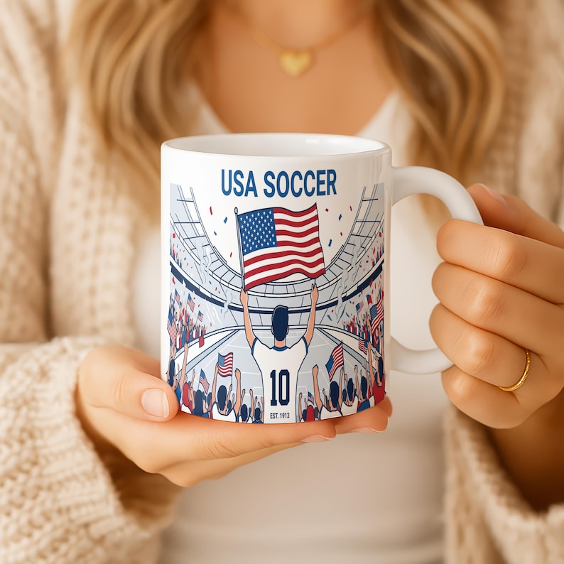 May include: White ceramic mug featuring a USA Soccer design. The mug displays the text "USA SOCCER" above an illustration of a soccer player holding an American flag in a stadium filled with fans. The player's jersey number is 10.