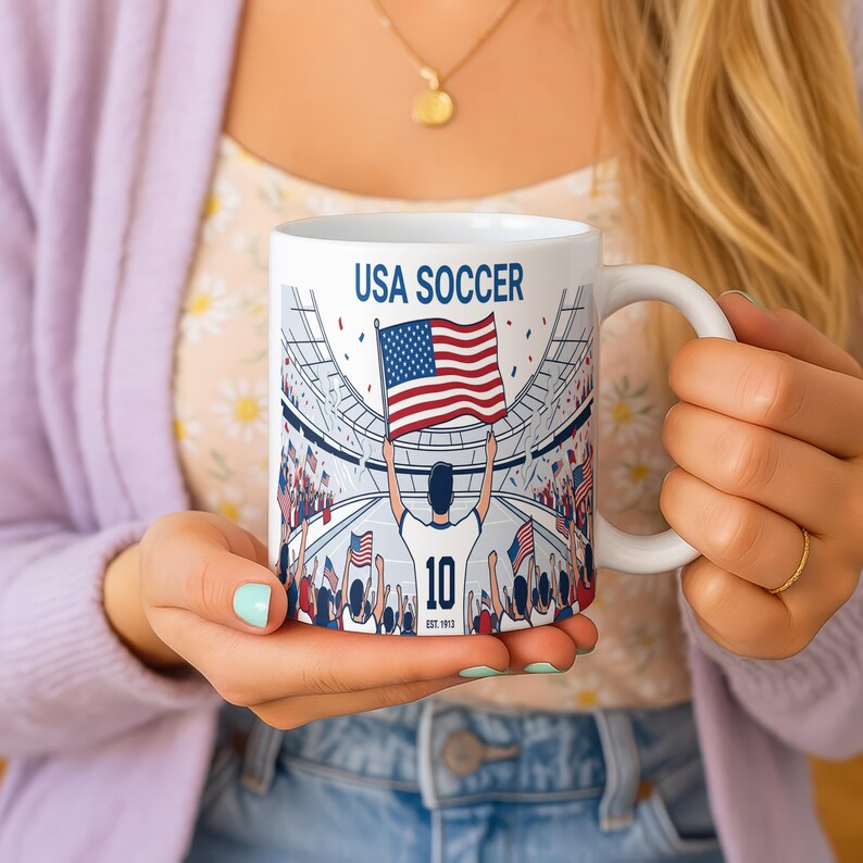 May include: White ceramic mug with the text "USA SOCCER" and a graphic of a soccer player holding the American flag in a stadium. The mug features a white handle and the number "10".