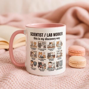 以下が含まれることがあります： ピンクの持ち手と縁が付いた白いセラミックマグ。テキスト「SCIENTIST / LAB WORKER this is my discovery way」と科学的プロセスを表す猫のイラストが特徴です。背景には2つのマカロンがあります。