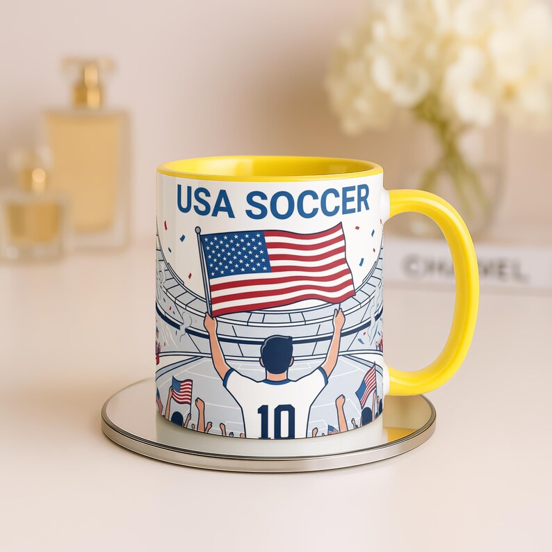 May include: A yellow-handled coffee mug with the words "USA SOCCER" and a graphic of a person holding an American flag in a stadium. The mug has a yellow interior and sits on a silver coaster.