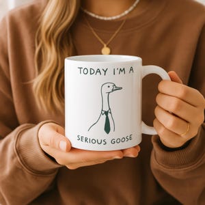 May include: White ceramic mug with the text "TODAY I'M A" above a cartoon goose wearing a tie, and the words "SERIOUS GOOSE" below. The mug has a curved handle and is held by a person.