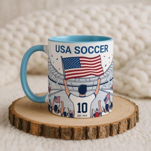 May include: White ceramic mug with a light blue handle and interior. The mug features a graphic of a soccer player holding an American flag in a stadium, with the text "USA SOCCER" and "10 EST. 1913".