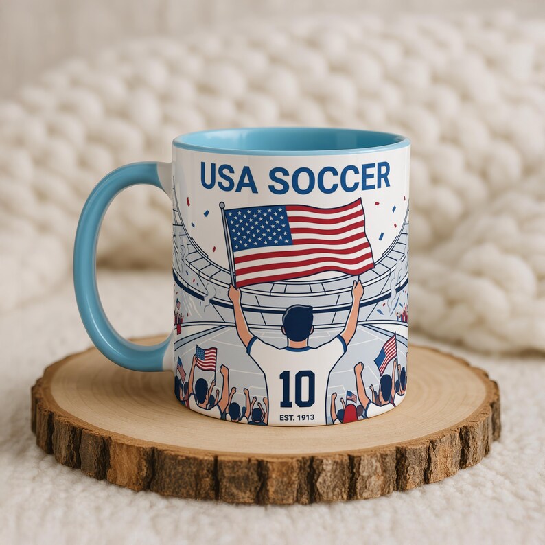May include: White ceramic mug with a light blue handle and interior. The mug features a graphic of a soccer player holding an American flag in a stadium, with the text "USA SOCCER" and "10 EST. 1913".