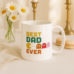 May include: White ceramic mug with the text "BEST DAD EVER" in yellow, green, and orange, along with Pac-Man-style graphics. The mug is on a white surface, with flowers and candles in the background.
