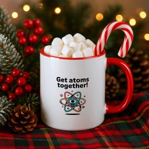 May include: A white ceramic mug with a red rim and handle, filled with white marshmallows and a red and white candy cane. The mug features the black text Get atoms together! above a colorful graphic of a cartoon atom with a smiling face.