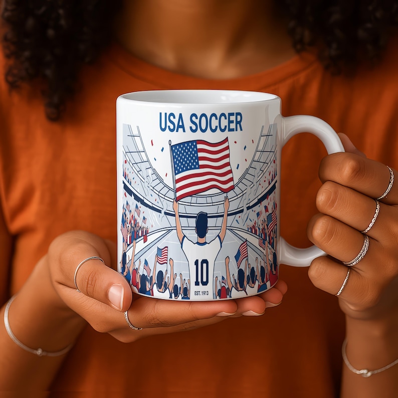 May include: White ceramic mug featuring a USA Soccer design. The mug displays a soccer player holding an American flag in a stadium setting, with the text "USA Soccer" and "10" visible. The mug has a white handle.