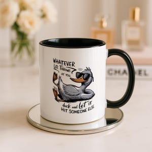 Könnte beinhalten: Ein weißer Keramikbecher mit schwarzem Innenraum und Henkel, verziert mit einer Grafik einer grauen Ente mit schwarzer Sonnenbrille. Der Text lautet: Whatever life throws at you, duck and let it hit someone else. Der Becher hat eine glatte Oberfläche.