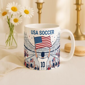 May include: White ceramic mug featuring a USA Soccer design. The mug displays a stadium scene with a person holding the American flag, the number 10, and the text "USA SOCCER". The mug is decorated with red, white, and blue colors.