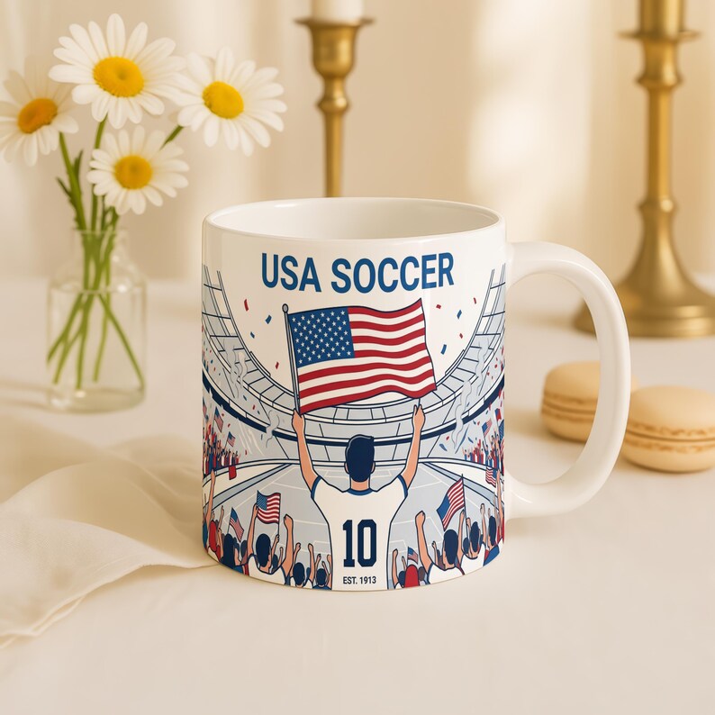 May include: White ceramic mug featuring a USA Soccer design. The mug displays a stadium scene with a person holding the American flag, the number 10, and the text "USA SOCCER". The mug is decorated with red, white, and blue colors.