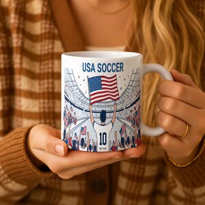 May include: White ceramic mug featuring a soccer stadium design with the text "USA SOCCER." The mug displays a player holding the American flag, with the number 10 and "EST. 1913" below. The design includes fans and confetti.