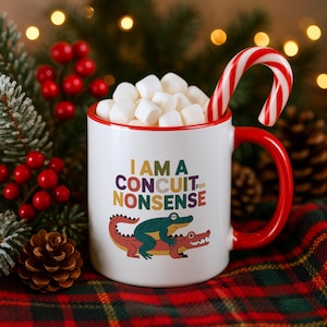 Peut inclure: Un mug en céramique blanc avec un bord et une anse rouges, rempli de guimauves blanches et d'un sucre d'orge rouge et blanc. Le mug porte le texte I AM A CONCUIT FOR NONSENSE au-dessus d'un crocodile vert chevauchant un crocodile rouge.