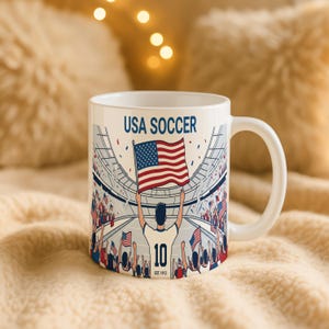 May include: White ceramic mug with a wraparound design featuring a soccer stadium scene. The design includes the text "USA SOCCER" and a player holding the American flag. The player's jersey has the number "10" and the text "EST. 1913".