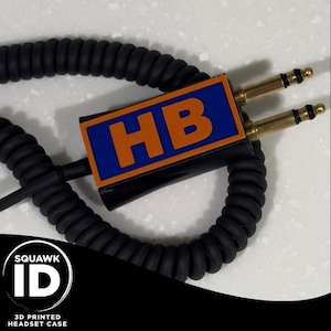May include: A black coiled cord with two gold-colored headphone jacks. The cord is attached to a black plastic case with a blue and orange label that says "HB".