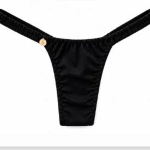 Puede incluir: Una braga de bikini tipo tanga de color negro con un diseño de tela fruncida a lo largo de la cintura y los laterales. La prenda incluye un pequeño dije de metal dorado de forma circular en el lado izquierdo con un logo S en relieve.