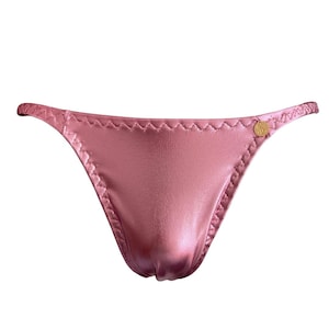 Könnte beinhalten: Rosa Satin-Unterwäsche mit String-Design. Die Kanten sind mit einer dekorativen Naht versehen. Auf der Seite befindet sich ein kleines goldfarbenes Emblem. Die Unterwäsche besteht aus einem glänzenden, glatten Stoff.