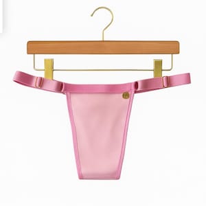 May include: A light pink thong with matching straps and gold-colored hardware. The lingerie is hanging on a wooden hanger with gold-colored metal accents. The thong has a satin finish.