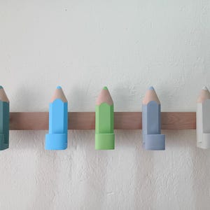 May include: Five wooden pencil-shaped coat hooks in different colors: light blue, green, gray, light blue, and gray. The hooks are mounted on a wooden board.