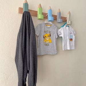 May include: A wooden wall-mounted coat rack with five pencil-shaped hooks. The hooks are painted in various colors, including blue, green, and light brown. A gray sweater, a gray t-shirt with the text "Play Eat Roar" and a white t-shirt are hanging on the hooks.