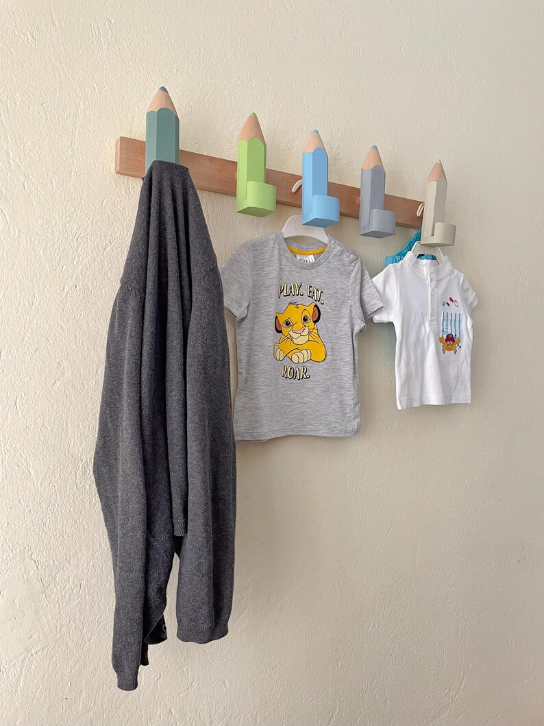 May include: A wooden wall-mounted coat rack with five pencil-shaped hooks. The hooks are painted in various colors, including blue, green, and light brown. A gray sweater, a gray t-shirt with the text "Play Eat Roar" and a white t-shirt are hanging on the hooks.
